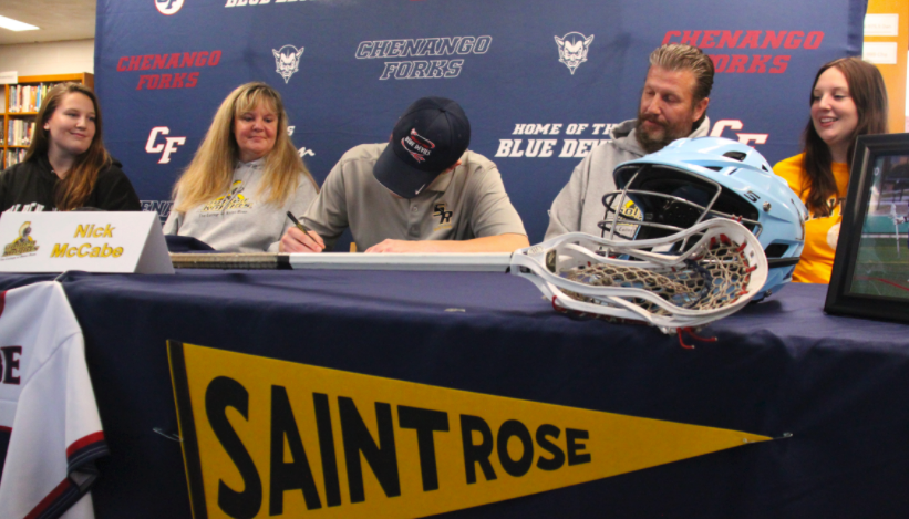 Nick McCabe signing NLI to play lacrosse at saint rose with a blue and yellow saint rose banner on table surrounded by family