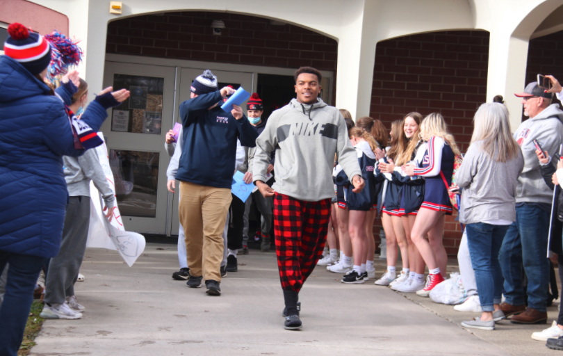 student in plaid pants and grey sweatshirt walking out of school building with staff and students cheering around him