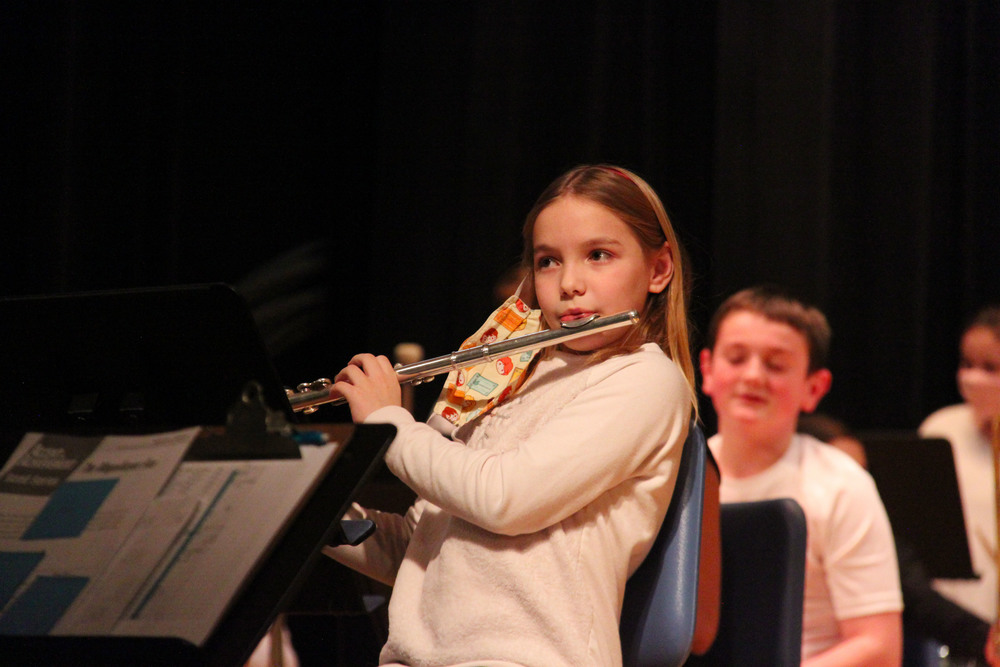 Young girl in white long sleeve shirt with blonde hair plays the flute at a band concert