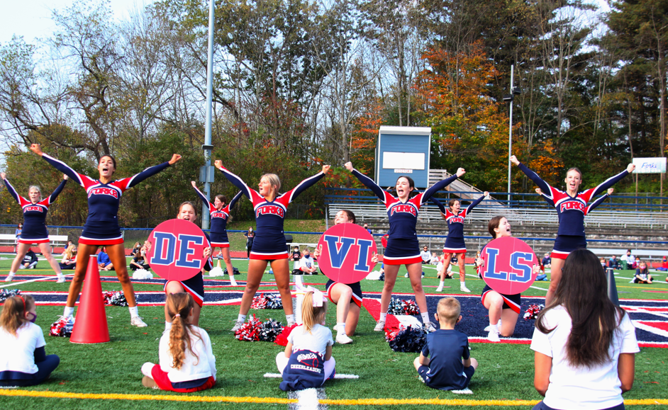 Cheerleaders in blue and red uniforms cheering on a football field in front of a group of students who are sitting